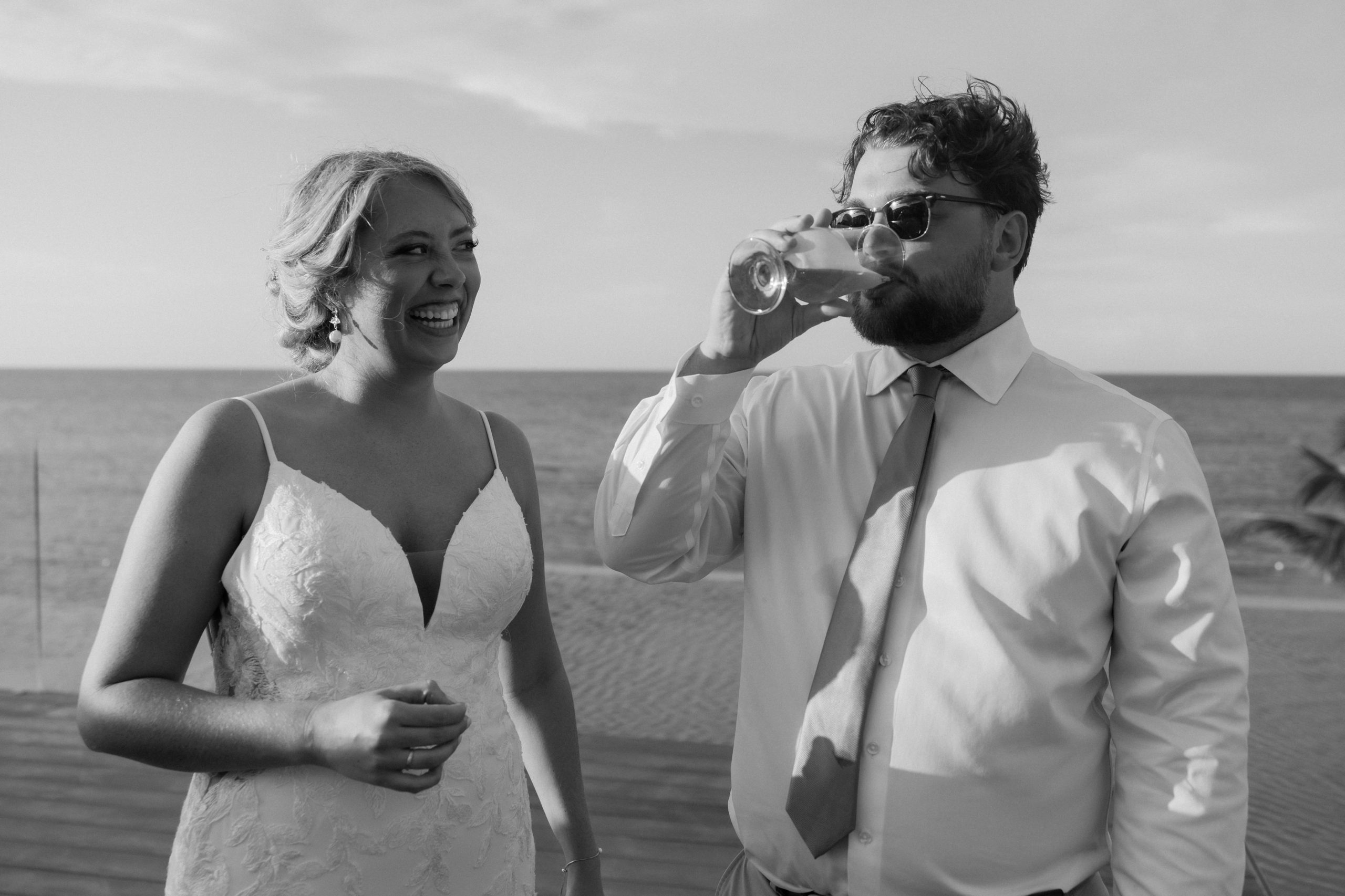 Groom drinks a cocktail while bride watches and smiles